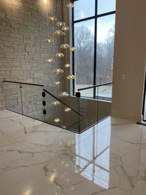 Frameless glass panels with a walnut handrail guard a stair opening on a glossy marble floor beneath a cascading glass pendant light.