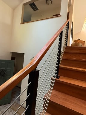 A wood handrail on black posts with stainless cables follows a cherry-toned stair.