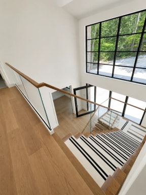 A glass guard with continuous oak handrail wraps a stairwell with covered treads beside large black-grid windows.