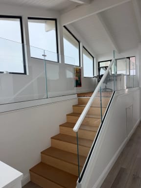Frameless glass with a slim white handrail follows an oak stair up to a glass-guarded loft below angled clerestory windows.