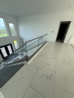 Frameless glass with a gray top cap rings a stair opening on a white marble-tile floor below tall windows.