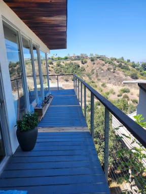 A narrow deck walkway with black-capped cable railing runs along a glass-walled home above dry canyon hills.