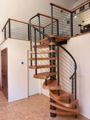 Light wood spiral stair on a black center column with curved cable railing up to a cable-railed loft.