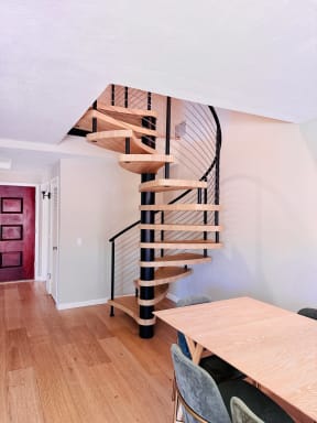 Light wood spiral stair on a black center column with curved cable railing beside a dining area.