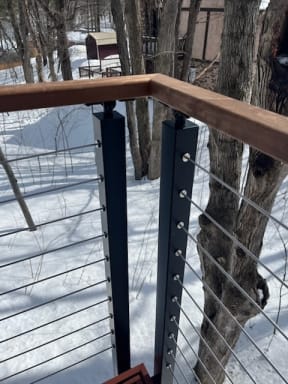 Two black slim side-mount posts at a deck corner with cable infill under a wood handrail.