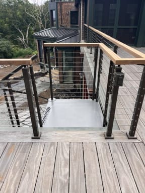 Black cable railing with wood cap frames a stair opening on a weathered wood deck.