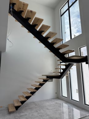 Black mono stringer with light wood treads climbs past tall windows over a polished stone floor.