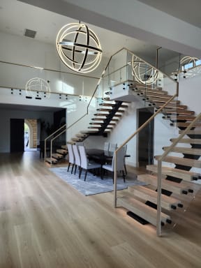 Pair of oak floating stairs on black mono stringers with frameless glass, wood handrails and LED glow framing a dining room under orb chandeliers.