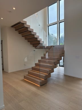 White oak stack stair with frameless glass railing and a glowing LED-lit bottom tread beside tall windows.