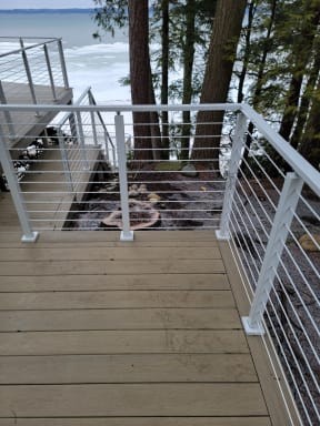 White posts with horizontal cable infill line a composite deck stair overlooking tall evergreens and water.