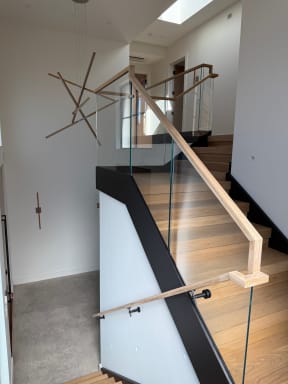 Frameless glass with a light wood handrail and a wall-mounted handrail line a wide oak stair under a sculptural light.