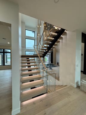 Light oak floating treads with under-tread LEDs on a black mono stringer rise alongside frameless glass in a bright modern stairwell.