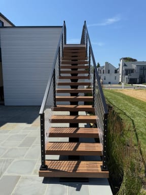 Straight-on view of slatted hardwood floating treads on a mono stringer rising between black cable railings from a bluestone patio.