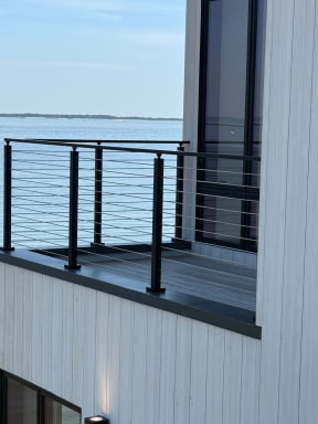 Black cable railing wraps a small balcony on a white-clad home overlooking open water.