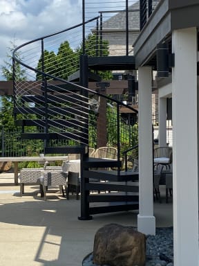 Black steel spiral stair with curved cable railing beside a white column and patio seating.