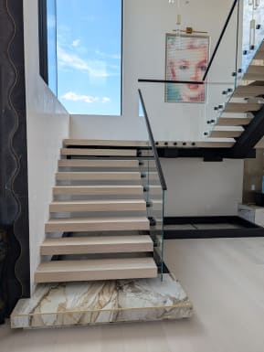 White oak floating treads rising from a gold-veined marble plinth with frameless glass and black handrail