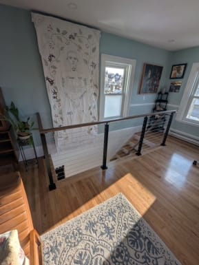 A black-post cable railing with wood handrail guards a stair opening in a sunlit living room with a patterned rug.