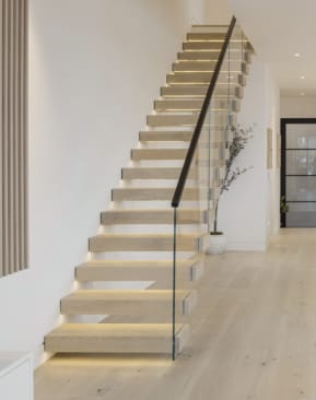 Light oak cantilever treads with under-tread LED lighting and a glass railing with black handrail rise along a white wall.