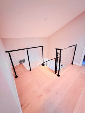 Black posts with cable infill guard a stair opening on light hardwood floors.