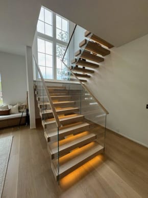 Light oak floating treads with warm under-tread LED lighting rise beside frameless glass with a wood handrail cap in a bright two-story room.