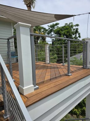 Silver-post cable railing wraps an IPE deck under a shade sail on a light green coastal home.
