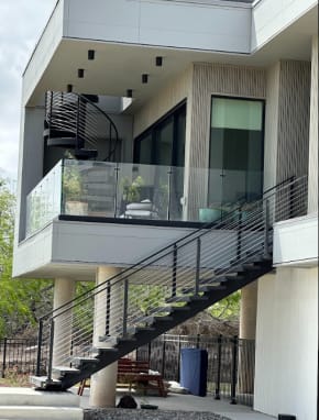 Elevated modern home with a black exterior stair with cable railing, glass balcony railing and spiral stair above.