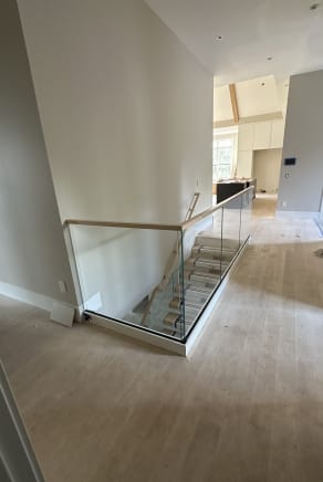 A frameless glass guard with oak handrail wraps an open stairwell in a bright hallway with light oak floors.