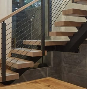 Light wood treads on a black steel mono stringer with a black cable railing against a board-formed concrete wall.