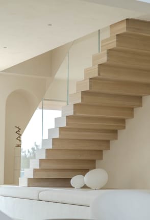 Light oak boxy stacked stair treads with no visible seams rise along a white wall beside a frameless glass railing.
