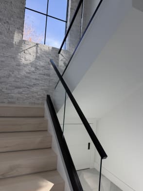 A white oak stair rises beside frameless glass on a kneewall with a black handrail toward a stone accent wall.