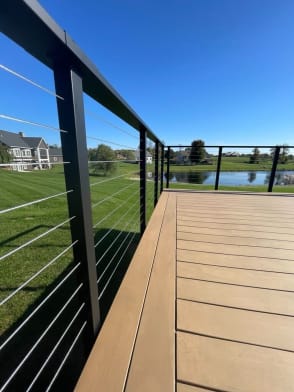 Black posts with cable infill and top rail along a composite deck overlooking a pond and lawn.
