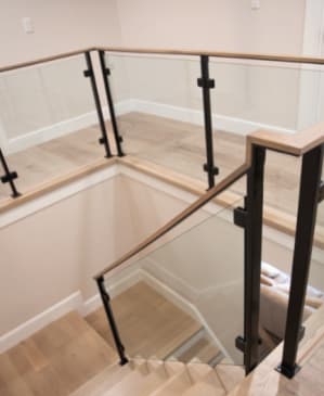 Black posts holding glass panels beneath a continuous light oak handrail wrapping a stair opening.