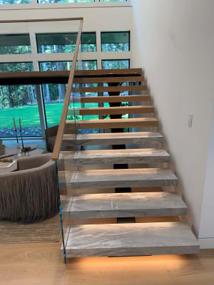 Thick marble-look floating treads glow with under-tread LEDs beside frameless glass and a wood handrail in front of tall windows.