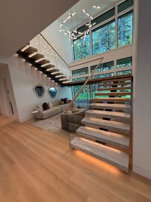 Floating stone-look treads with LED lighting and glass railing rise along a wall of windows in a vaulted living room.