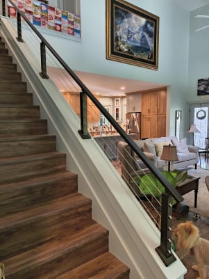 Burnished bronze cable railing on a white knee wall follows a wood stair beside an open living room.