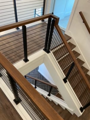 Black posts with thick black horizontal rods and a wood handrail top a white kneewall around a switchback stairwell.