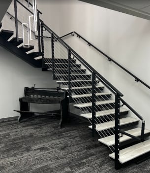 Black mono stringer switchback stair with gray wood-look treads, black cable railing and wall handrail in an office lobby.