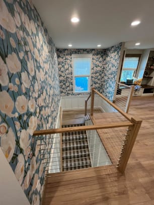 Oak newel posts and cap rail with stainless cable infill around a carpeted stair opening with floral wallpaper.