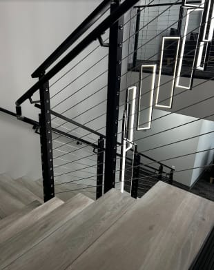 Black cable railing with wall handrail along a switchback stair under hanging rectangular pendant lights.