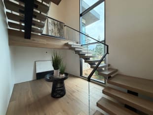 Black mono-stringer stair with thick oak treads and glass railing turns past a tall window.