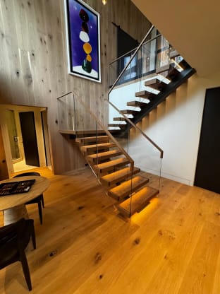 Thick oak treads with warm under-tread LED lighting float on black stringers beside frameless glass with a wood handrail in a double-height wood-paneled room.