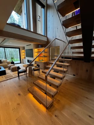 Oak floating treads glowing with under-tread LED light climb past a frameless glass guard with a wood handrail into an open living room.