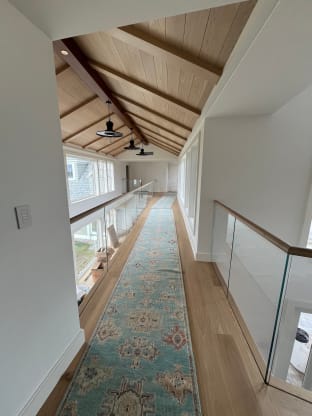 Frameless glass railing with a white oak cap rail lines a long vaulted upstairs hallway with a runner rug.