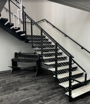 Black mono stringer switchback stair with gray wood-look treads, black cable railing and wall handrail in an office lobby.