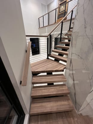 Wood floating treads turn on a black mono stringer with cable railing beside a marble wall.