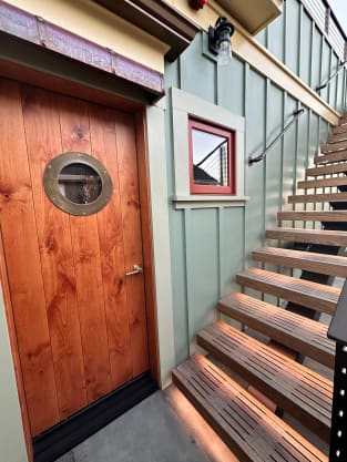 Lit slatted exterior treads rise beside a rustic wood door with a porthole window on a green-sided home.