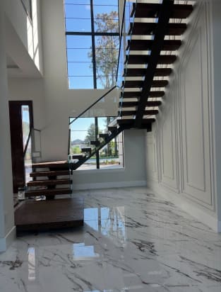 Dark floating treads on a black mono stringer rise from plinths with glass railing above a white marble floor.