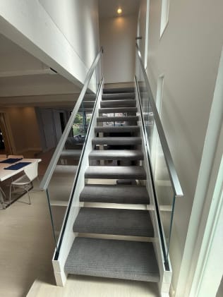 Narrow carpeted stair flanked by frameless glass in a white base rail channel with stainless handrails.