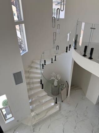 Curved glass panels on black talon spigots follow a sweeping white marble stair beneath a crystal chandelier.