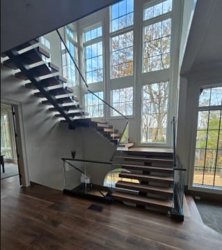 Walnut treads on a black mono stringer with frameless glass in a base rail beneath a two-story window wall.